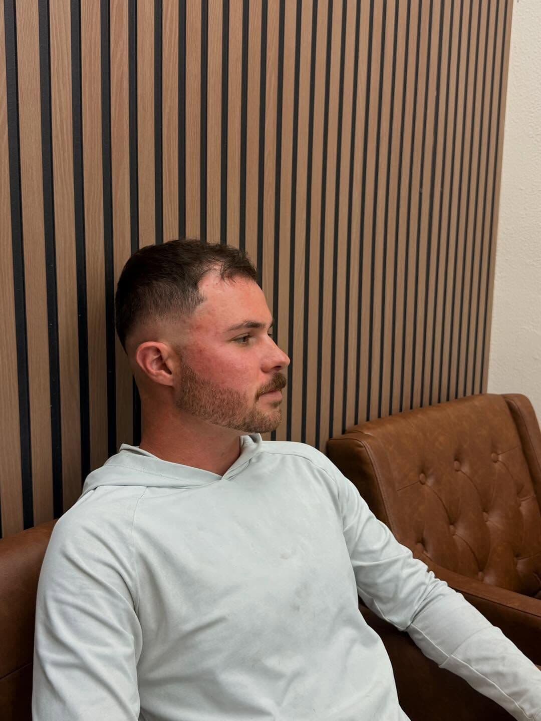 A client with a high fade and a short beard, seated in a leather chair by the wood-slat wall