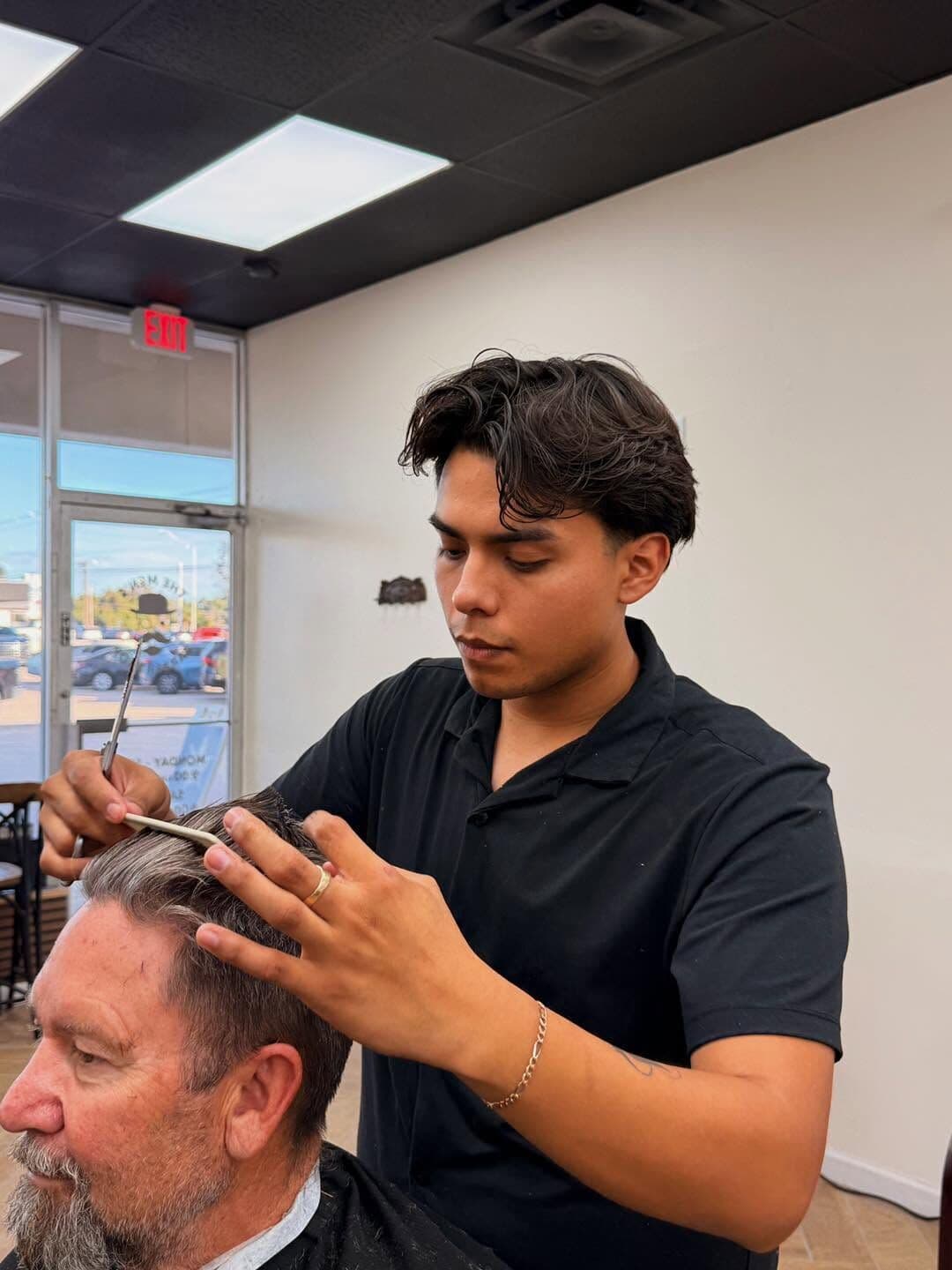 Moises cutting a client's gray hair with scissors and a comb, by the front window