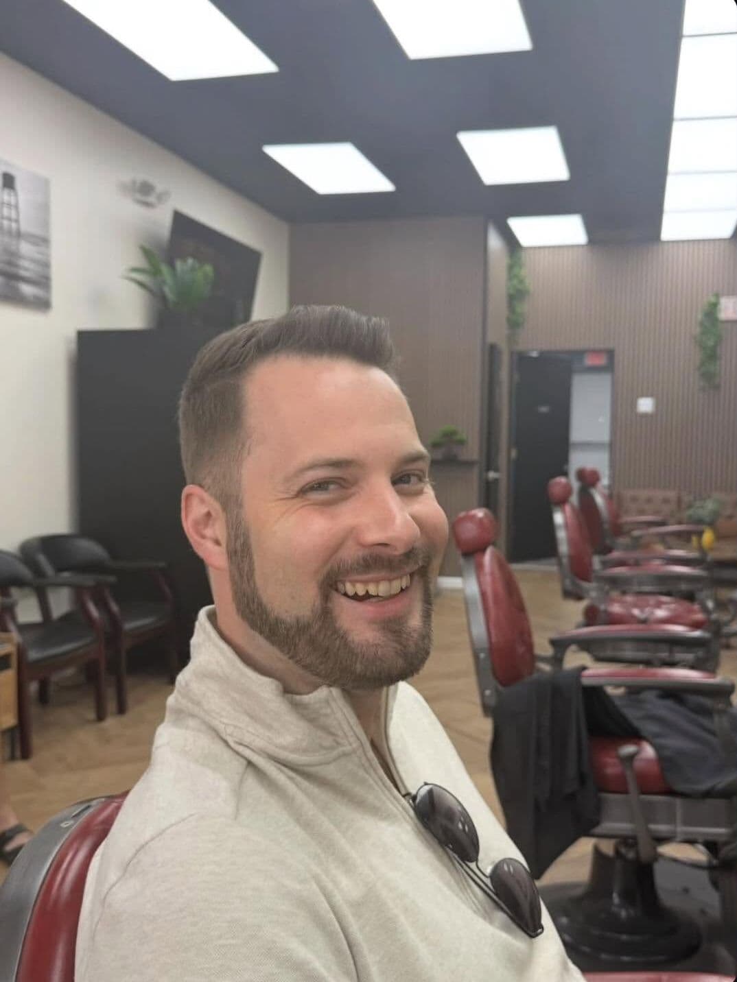 A smiling client with a short side part and a shaped beard, in one of the red barber chairs