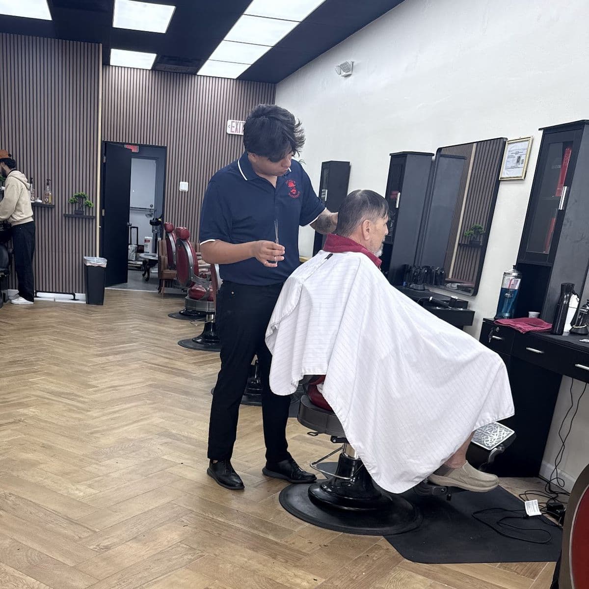 A barber cutting a client's hair in a red barber chair, with the stations behind